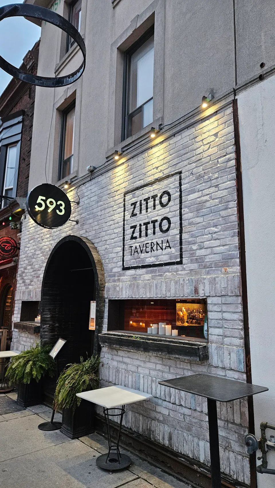 The exterior of Zitto Zitto Taverna, featuring a white brick facade, an arched doorway, outdoor tables, and a sign with the restaurant's name and address number 593.
