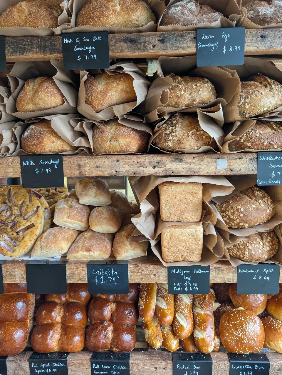 A rustic wooden bakery shelf filled with various freshly baked artisanal breads, including sourdough loaves, ciabatta, multigrain pullman, and pretzel buns, each with handwritten price tags.