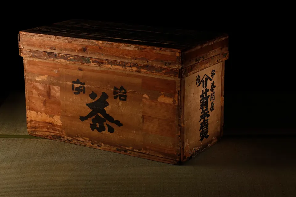 An antique wooden tea chest with Japanese calligraphy characters painted on its side, resting on a traditional tatami mat under dramatic, low-key lighting.