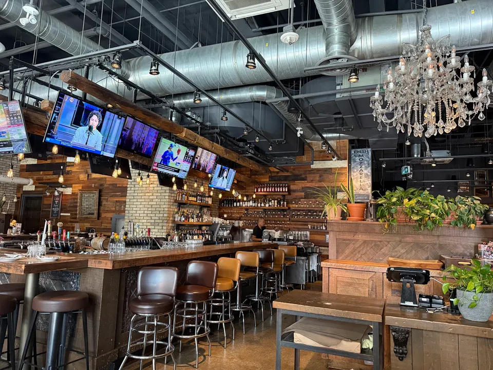A rustic bar area with wooden paneling, brick accents, leather bar stools, multiple overhead TV screens, and a large crystal chandelier hanging from an industrial ceiling.