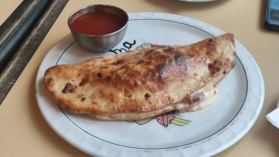 A golden-brown baked calzone oozing with cheese and fillings, served on a white plate with a small metal bowl of red marinara sauce on the side.