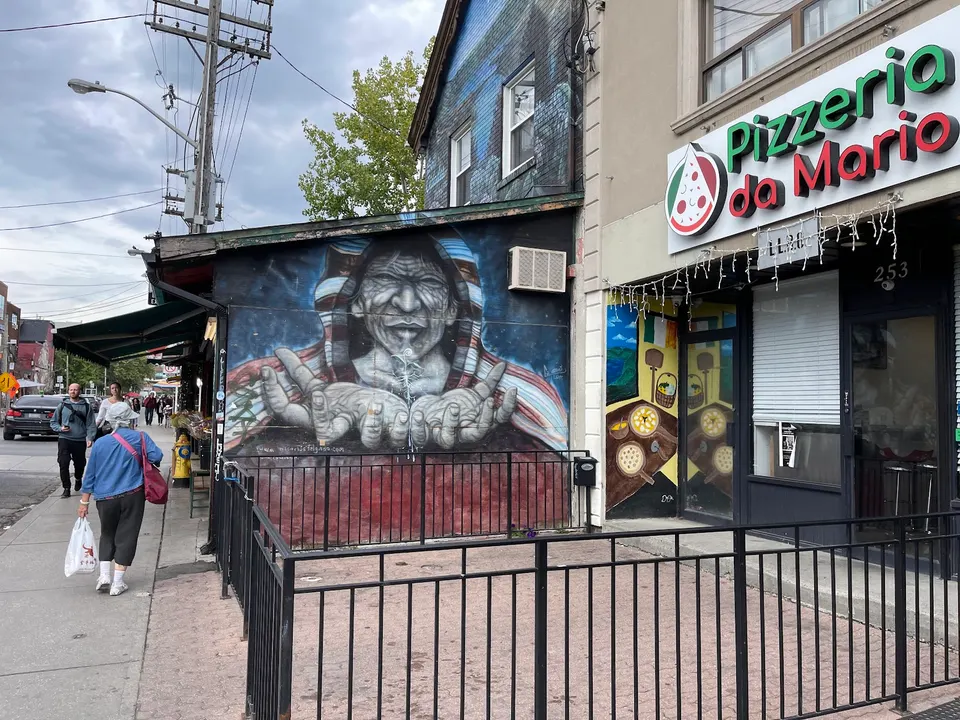 Exterior view of Pizzeria da Mario in Kensington, featuring a green and red sign, storefront windows, and a large colorful mural of a face on the adjacent brick wall.