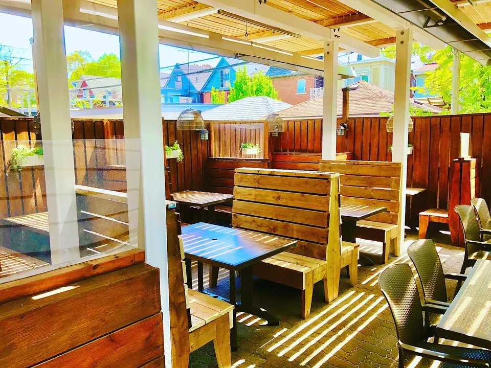 An outdoor patio dining area with wooden booths, tables, and dark wicker chairs under a covered wooden pergola structure.