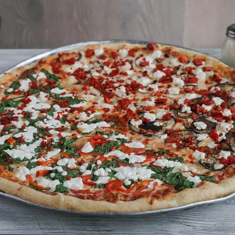A large, thin-crust pizza divided into four distinct quarters with toppings including spinach, red peppers, mushrooms, eggplant, sun-dried tomatoes, and crumbled goat cheese.