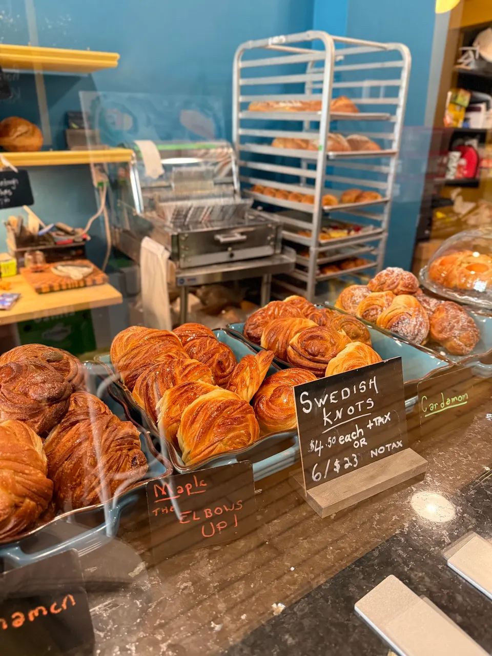 A bakery display counter showcasing rows of golden-brown Swedish knots in blue ceramic dishes, with handwritten price signs and a tall metal baking rack in the background.