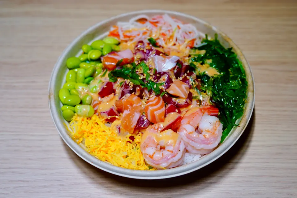 A colorful poke bowl in a round paper container filled with salmon, tuna, shrimp, edamame, seaweed salad, shredded egg, and crab salad, drizzled with spicy mayo.