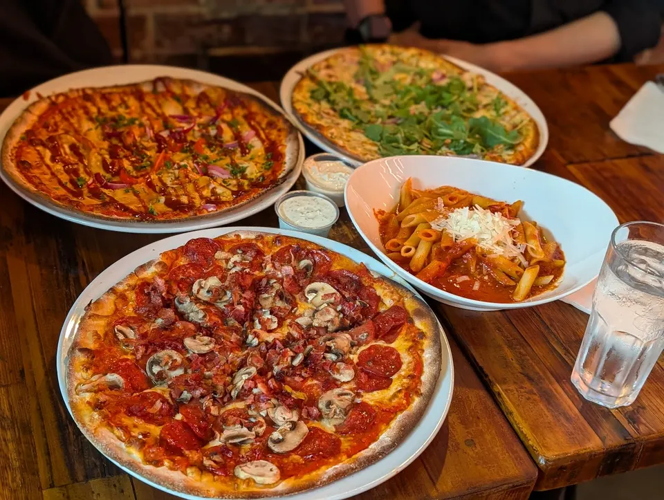 A wooden table spread with three different pizzas, a bowl of penne pasta with red sauce and cheese, two small dipping sauce cups, and a glass of water.