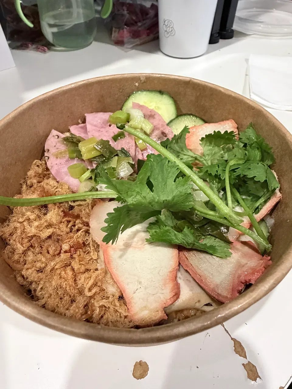 A Vietnamese noodle or rice bowl in a cardboard container, topped with sliced pork, Vietnamese ham, pork floss, cucumber slices, and fresh cilantro.