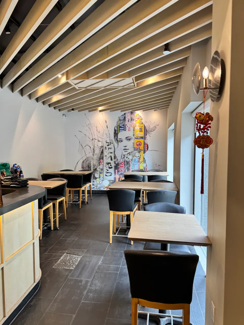 An empty dining area at KAKA All You Can Eat featuring wooden tables, black high-top chairs, a slatted wooden ceiling, and a large wall mural of a woman's face in a modern artistic style.