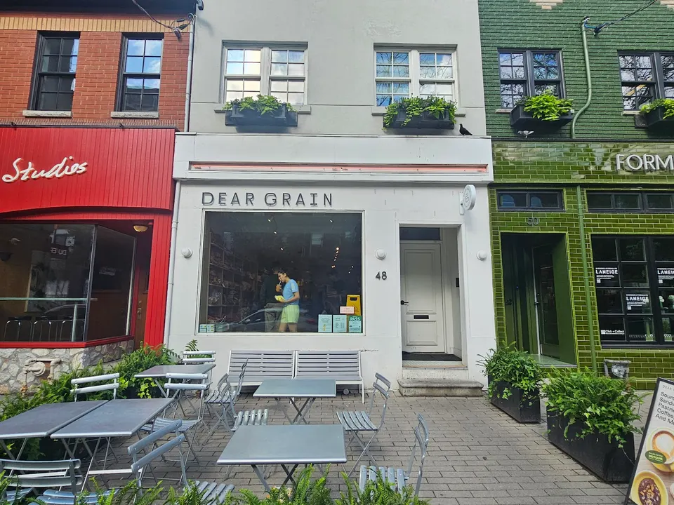 The storefront of Dear Grain bakery, featuring a white facade, large window showing the interior, outdoor patio tables and chairs, and a sign reading 'DEAR GRAIN'.