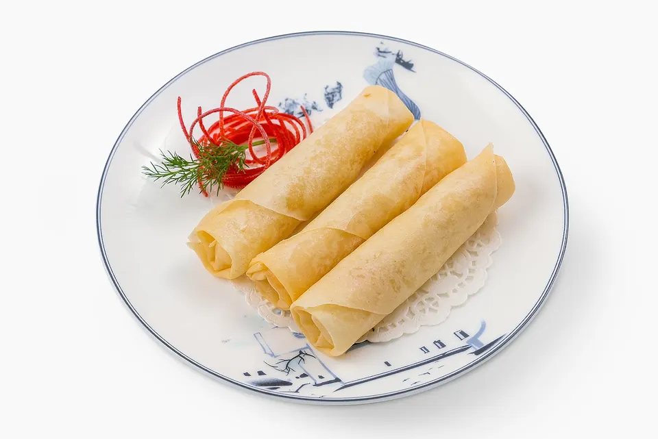 Three golden-brown spring rolls arranged diagonally on a white plate with blue patterns, garnished with a sprig of dill and red curled ribbons.