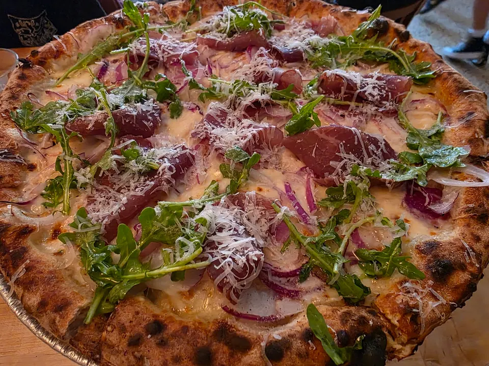 A freshly baked gourmet pizza with a charred crust, topped with melted white cheese, red onions, cured meat slices, fresh arugula, and grated parmesan cheese.