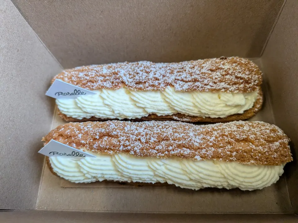 Two long, cream-filled eclairs dusted with powdered sugar, each with a small white Roselle paper tag, sitting side-by-side inside a cardboard takeout box.