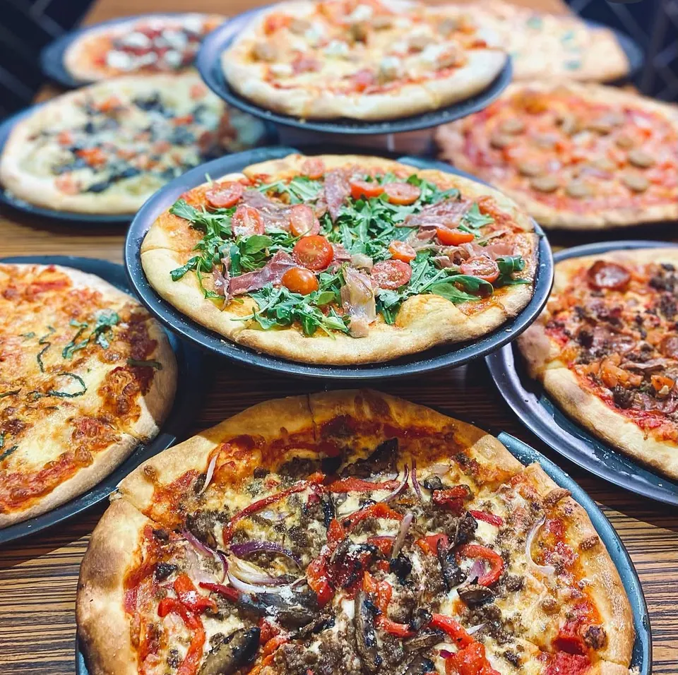A table filled with several freshly baked artisanal pizzas on black plates, featuring a variety of toppings like arugula and prosciutto, mushrooms and peppers, and classic pepperoni.