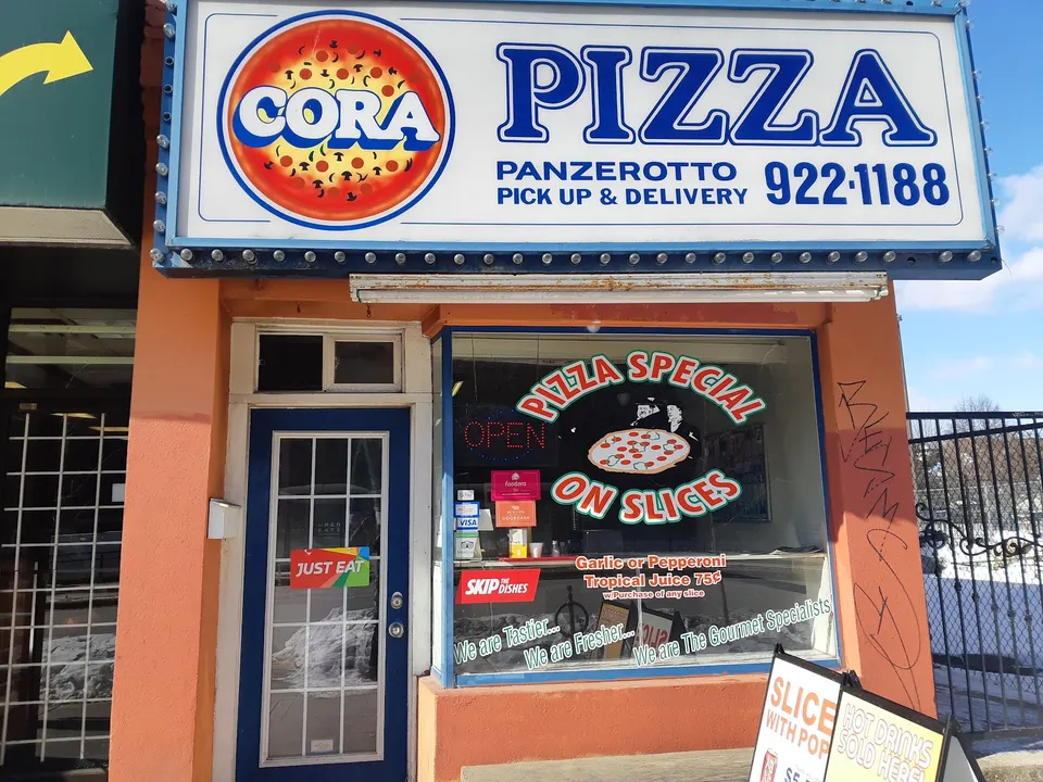 Exterior storefront of Cora Pizza featuring a large blue and white sign with a pizza graphic, orange painted walls, and a window displaying pizza specials.