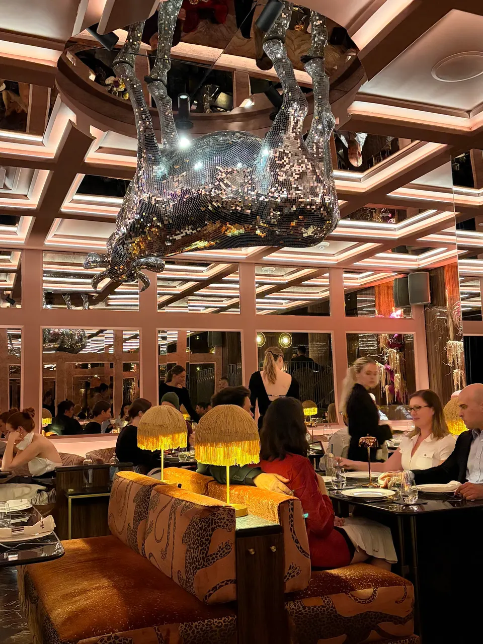 An upscale restaurant dining room featuring a large, sparkling disco-ball style cow sculpture hanging upside down from a mirrored ceiling above patrons seated at tables.