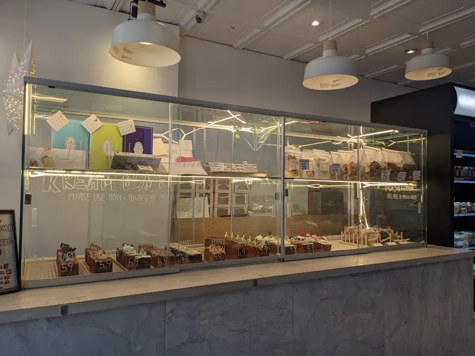 A glass display case at Kream bakery filled with various pastries, cakes, and packaged treats, set against a marble counter under warm hanging lights.