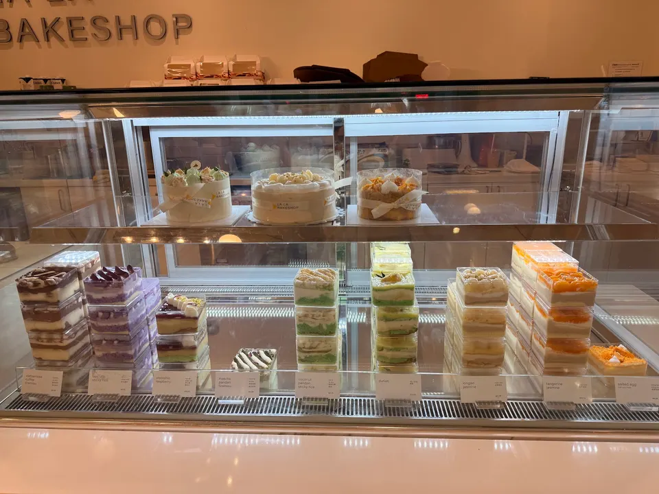 A glass display case at LA LA Bakeshop filled with various cakes, including whole decorated cakes on the top shelf and individual rectangular cake slices stacked on the bottom shelf.