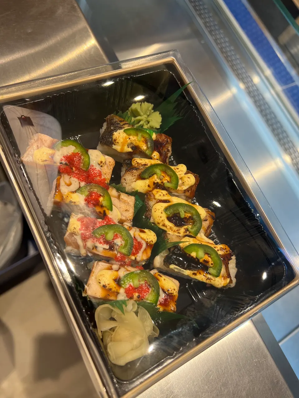 A black plastic takeout container filled with eight pieces of seared sushi rolls topped with sliced jalapeños, creamy sauce, and red fish roe, served with ginger and wasabi.
