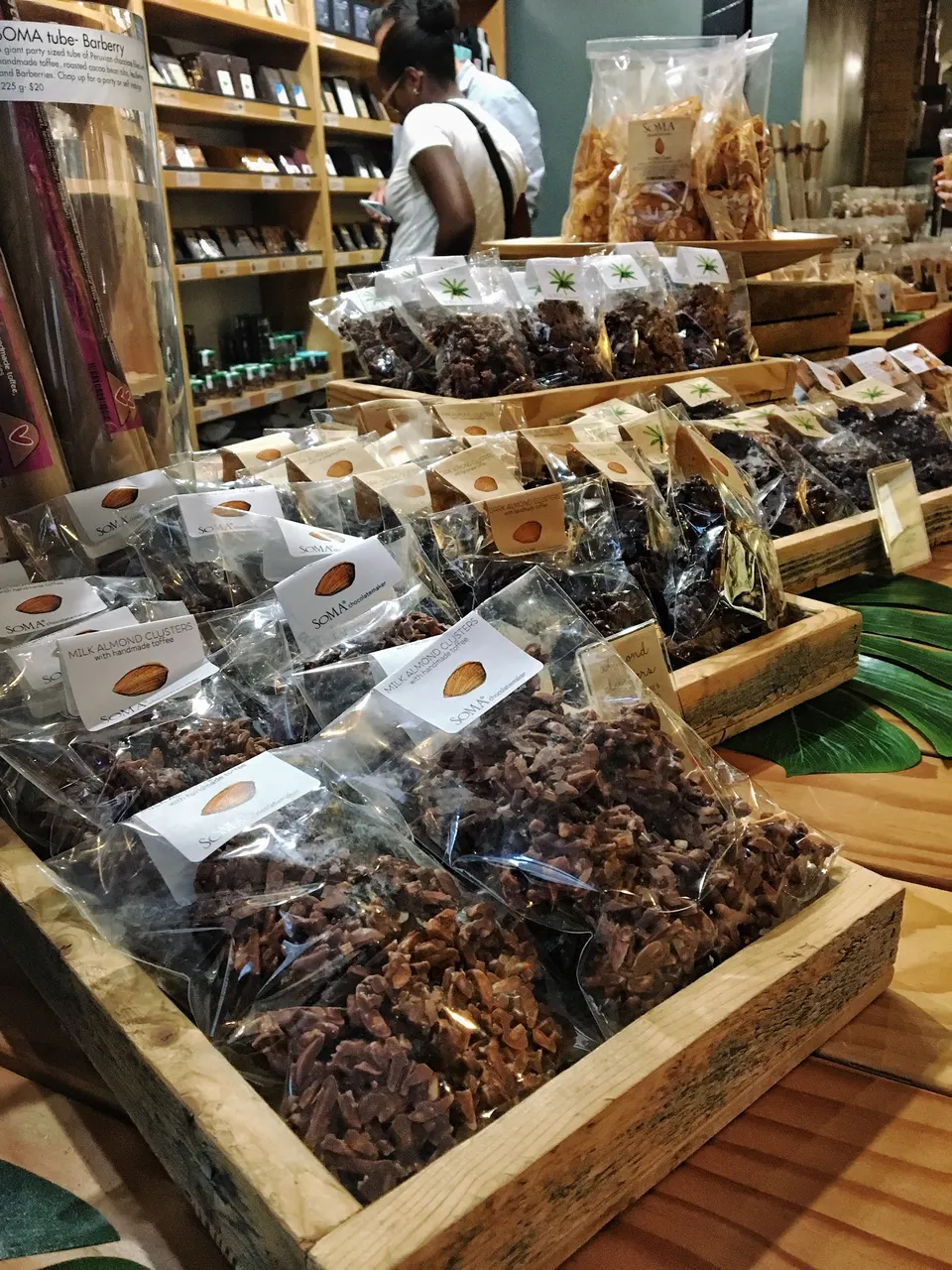 Wooden trays filled with packaged chocolate almond clusters on display at SOMA Chocolatemaker, with shelves of chocolate bars and customers in the background.