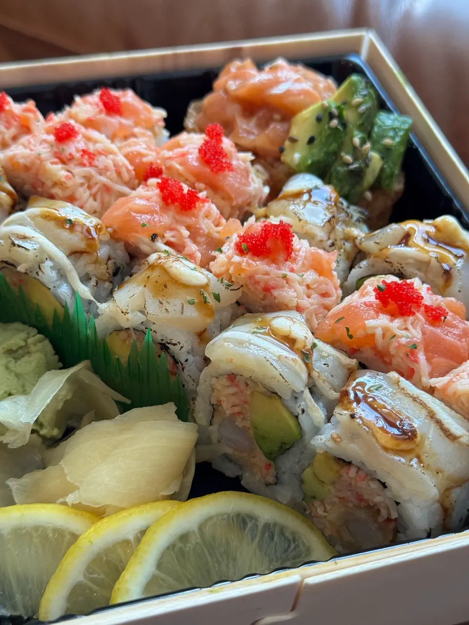 A close-up view of a sushi platter featuring various rolls topped with salmon, crab salad, avocado, and red tobiko, served with pickled ginger, wasabi, and lemon slices in a wooden box.