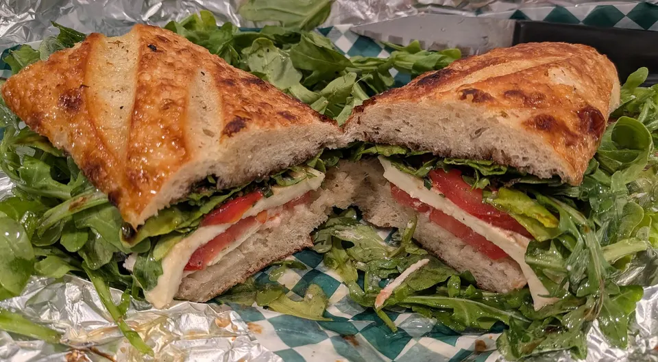 A caprese sandwich cut in half on a baguette with fresh mozzarella, sliced tomatoes, and basil, served over a bed of fresh arugula on checkered paper.