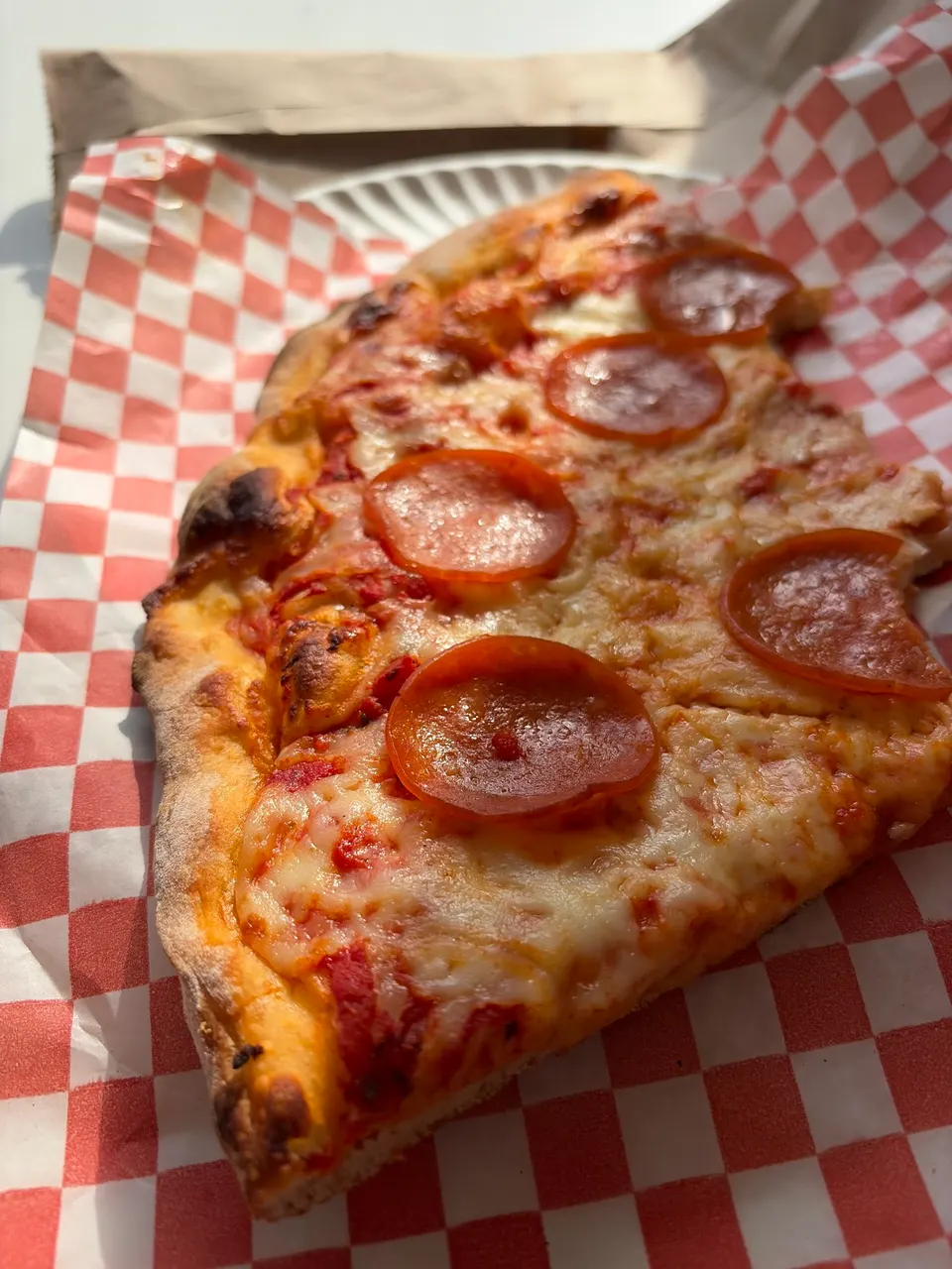 A thick slice of pepperoni pizza with melted cheese and tomato sauce on red and white checkered paper.