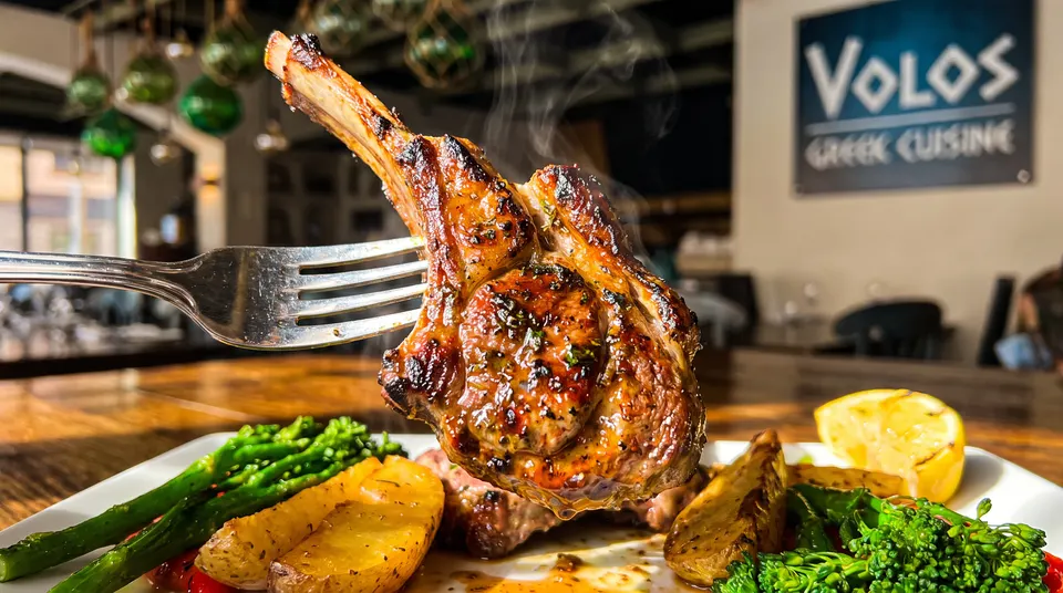 A grilled lamb chop held up on a fork, showing steam rising from the meat, with roasted potatoes, broccolini, and a lemon wedge on a plate below, set against a blurred restaurant background with a Volos Greek Cuisine sign.