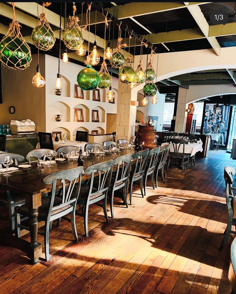 A long wooden dining table set with wine glasses and plates under hanging green glass buoy lights and exposed bulbs in a rustic Greek restaurant with hardwood floors.
