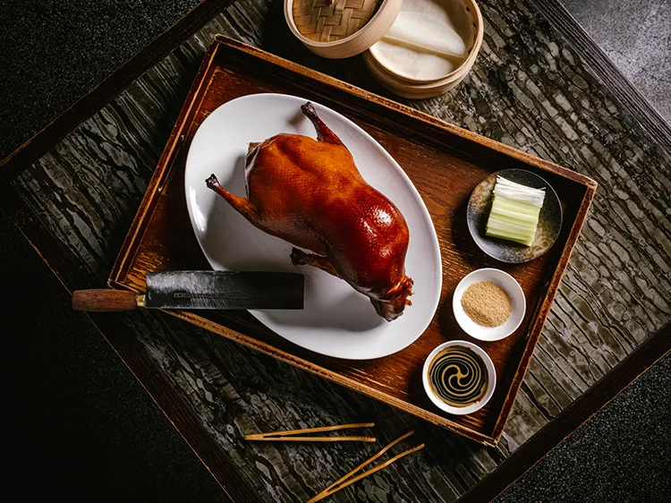 A whole roasted Peking duck with glossy, crispy skin presented on a white platter on a wooden tray, accompanied by sliced scallions, sugar, hoisin sauce, thin pancakes in a bamboo steamer, and a cleaver.
