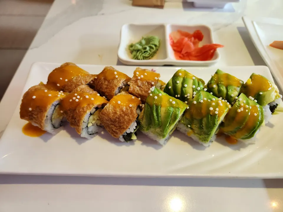 A white rectangular plate of sushi rolls topped with mango sauce and tiny white pearls, featuring half avocado-topped rolls and half inari-wrapped rolls, served with wasabi and pickled ginger.