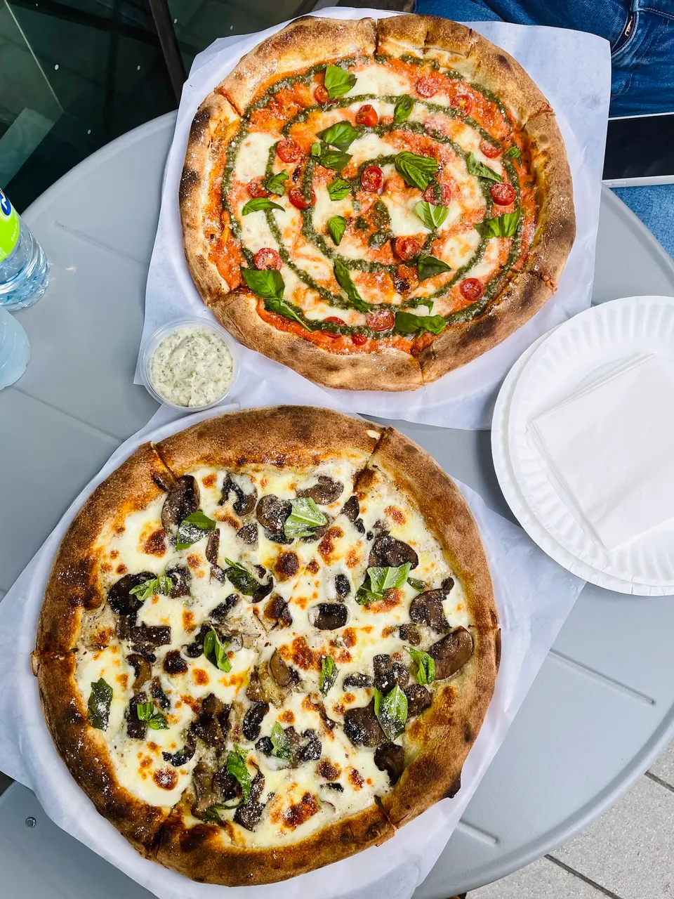 Two whole pizzas served on paper liners on a grey outdoor table, one topped with red sauce, pesto swirl, cherry tomatoes, and fresh basil, and the other with white sauce, mushrooms, and basil.