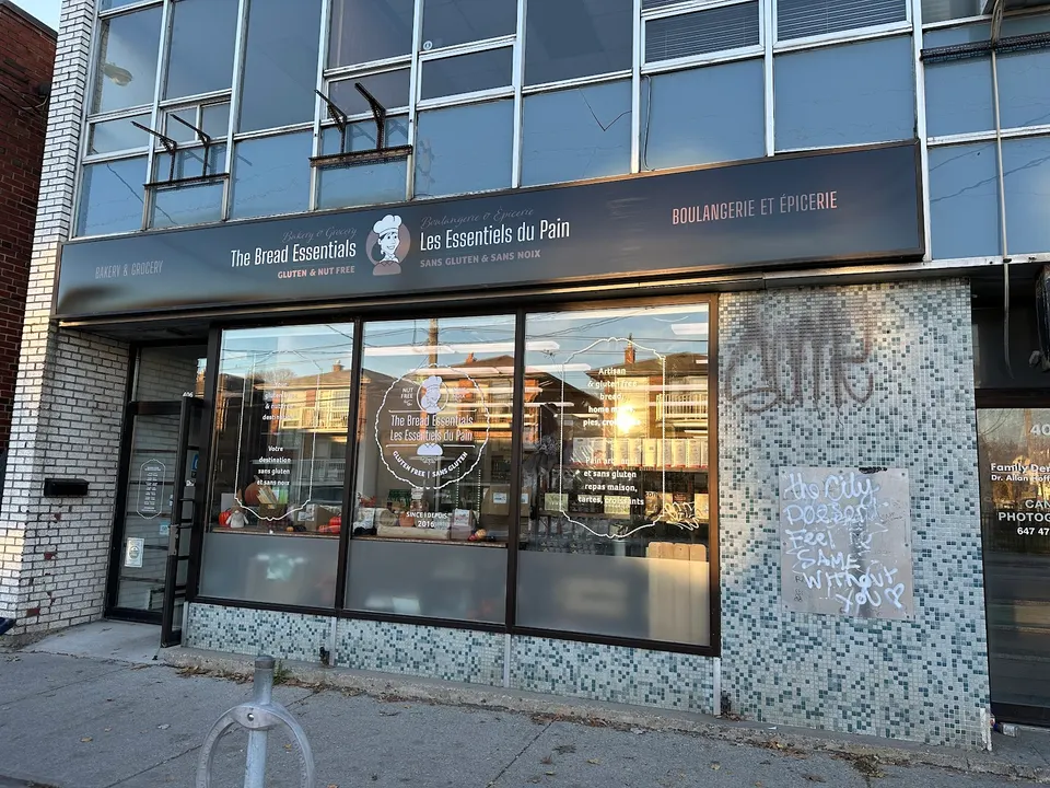 The storefront of The Bread Essentials bakery and grocery, featuring a dark sign with English and French text, large glass windows displaying products, and a mosaic tile wall with graffiti.