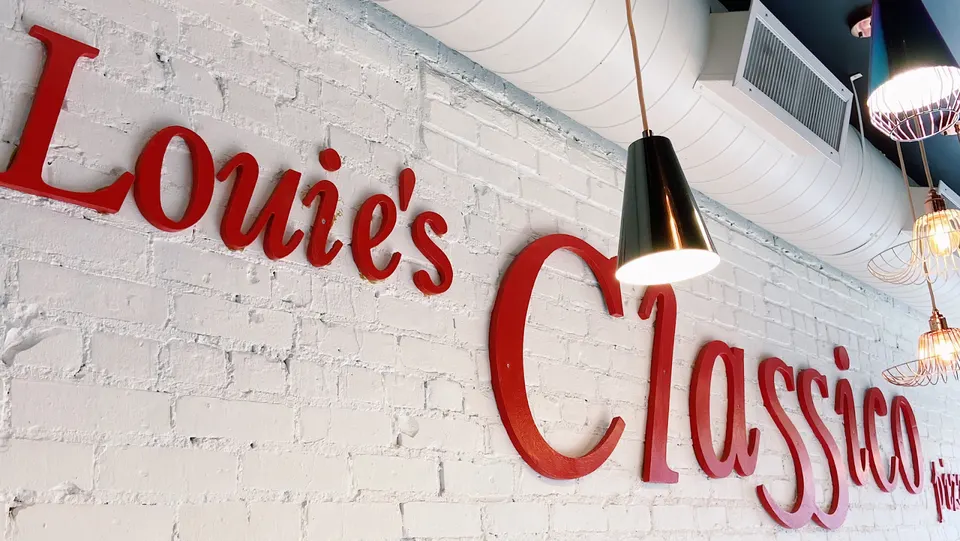 Red dimensional lettering spelling out 'Louie's Classico' mounted on a white painted brick wall inside a restaurant, illuminated by hanging pendant lights.