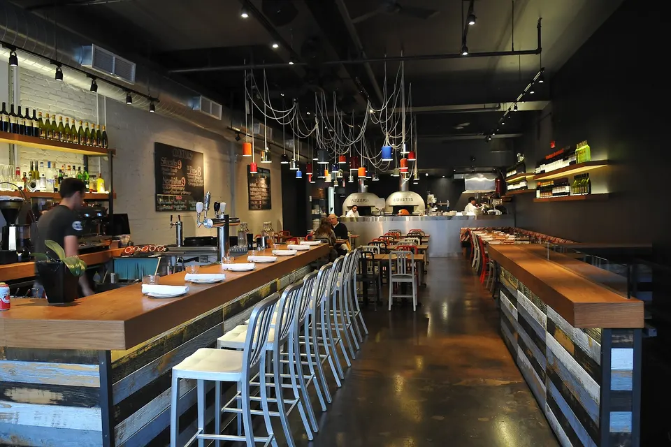 Interior of a modern pizzeria featuring a long wooden bar with metal stools, dining tables in the background, and a view of the open kitchen with pizza ovens.