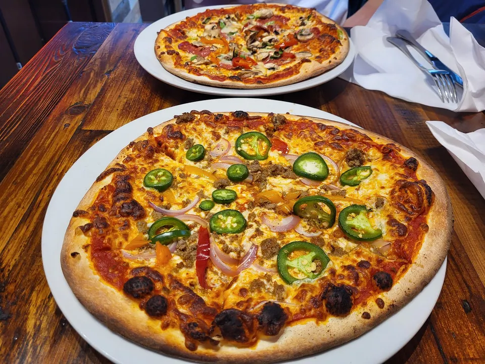 Two freshly baked thin-crust pizzas served on white plates on a rustic wooden table. The foreground pizza is topped with jalapeños, red onions, and ground meat.