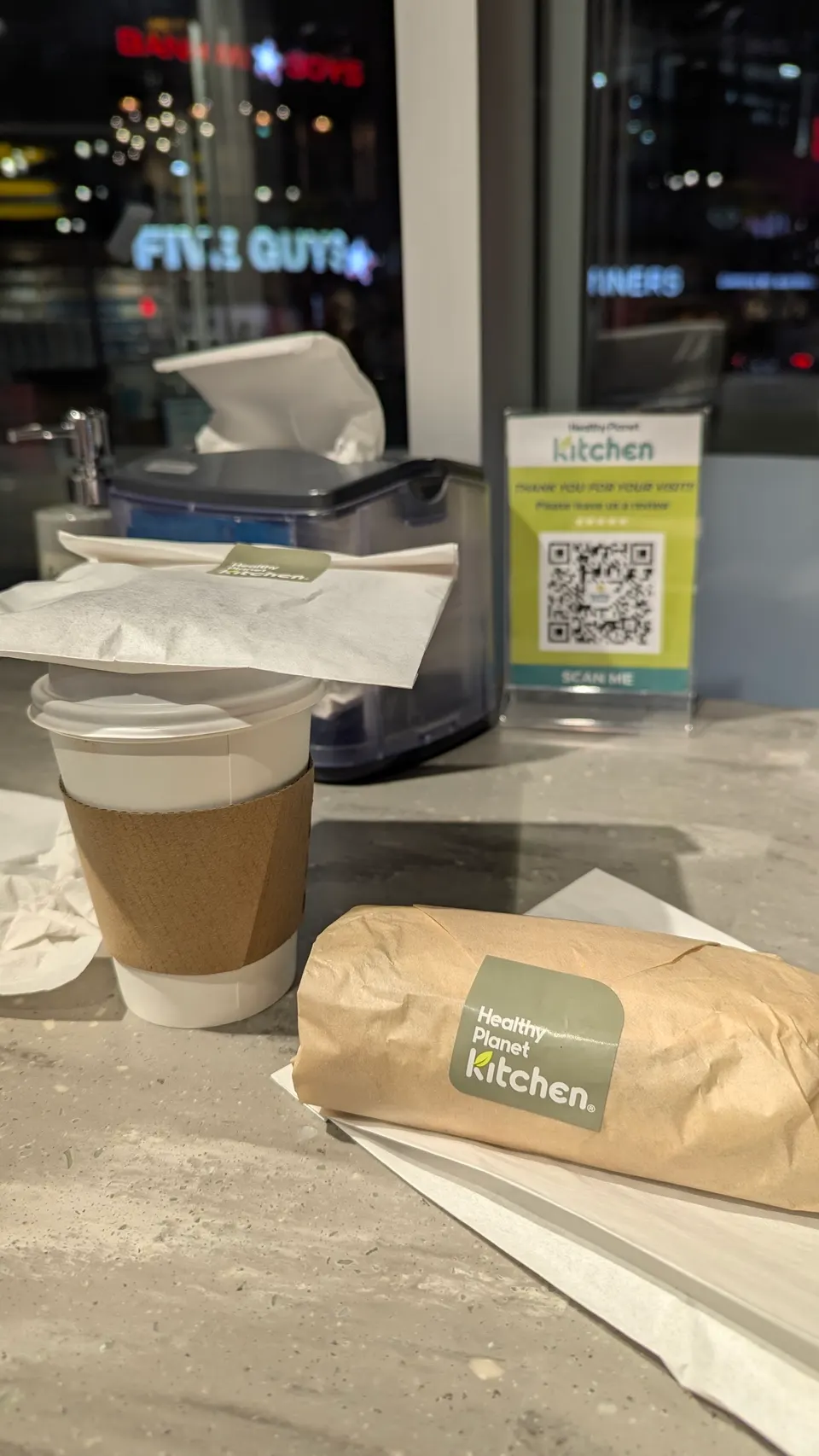 A wrapped sandwich with a Healthy Planet Kitchen sticker sits on a napkin next to a hot beverage in a paper cup with a cardboard sleeve, set on a counter.
