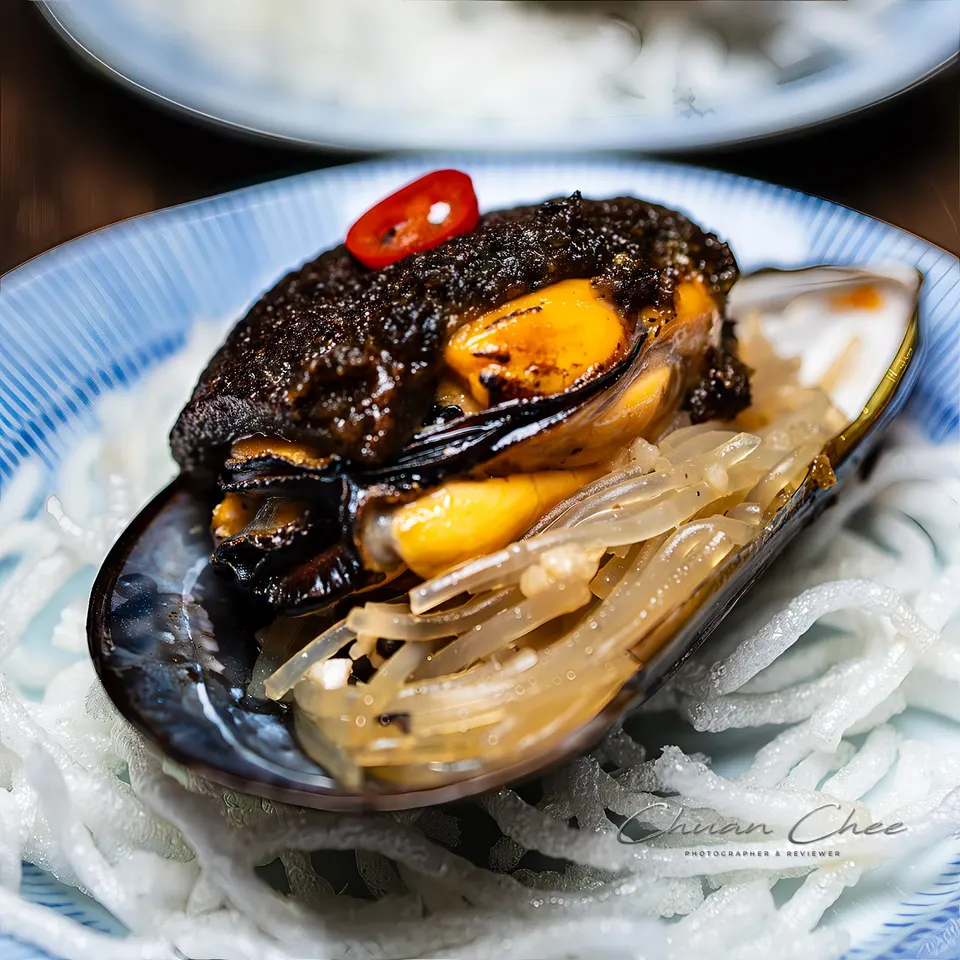 A single cooked mussel served in its shell, topped with a dark savory paste and a slice of red chili, resting on a bed of translucent noodles inside a blue and white patterned dish.