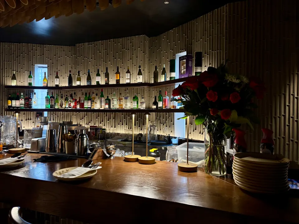 A dimly lit bar counter at TRE - Viet Heritage Kitchen & Bar, featuring shelves of liquor bottles against a textured bamboo-style wall, small table lamps, a vase of red roses, and stacked plates.