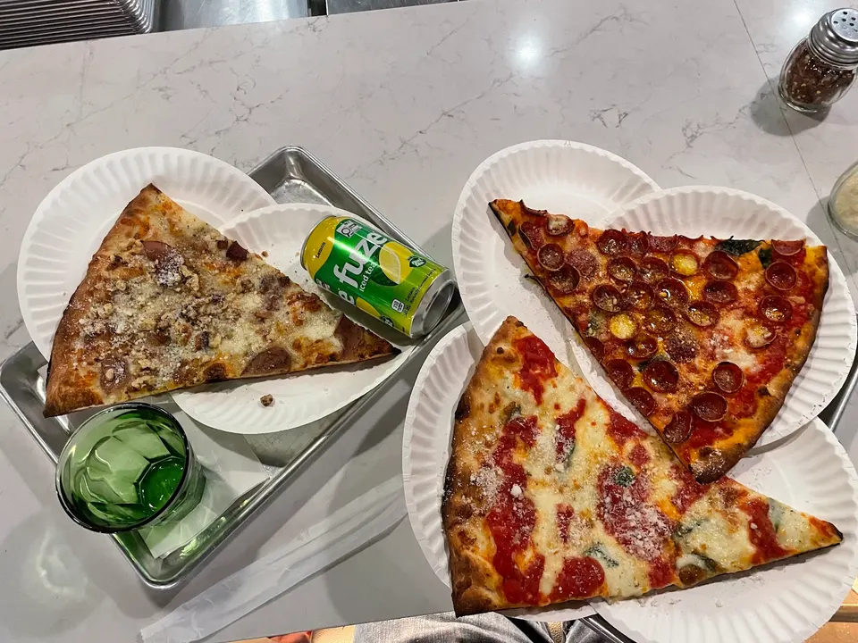 Three large slices of thin-crust pizza on paper plates, including pepperoni, margherita, and a white sauce slice, served on a metal tray with a can of Fuze iced tea and a green glass.