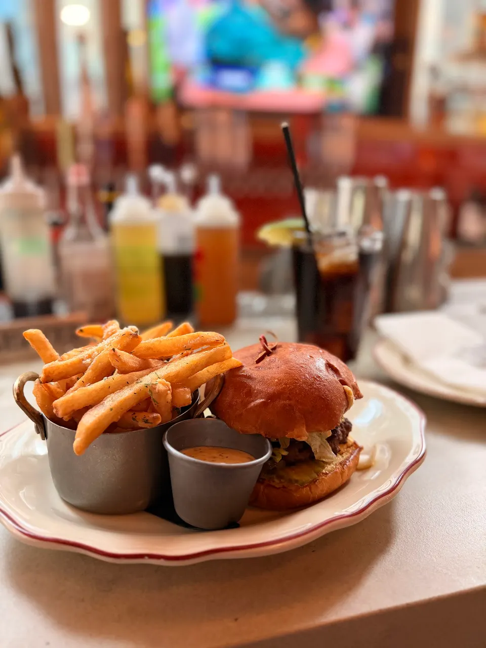A gourmet burger on a brioche bun served with a metal cup of seasoned french fries and a small ramekin of dipping sauce on a white plate, set against a blurred bar background.