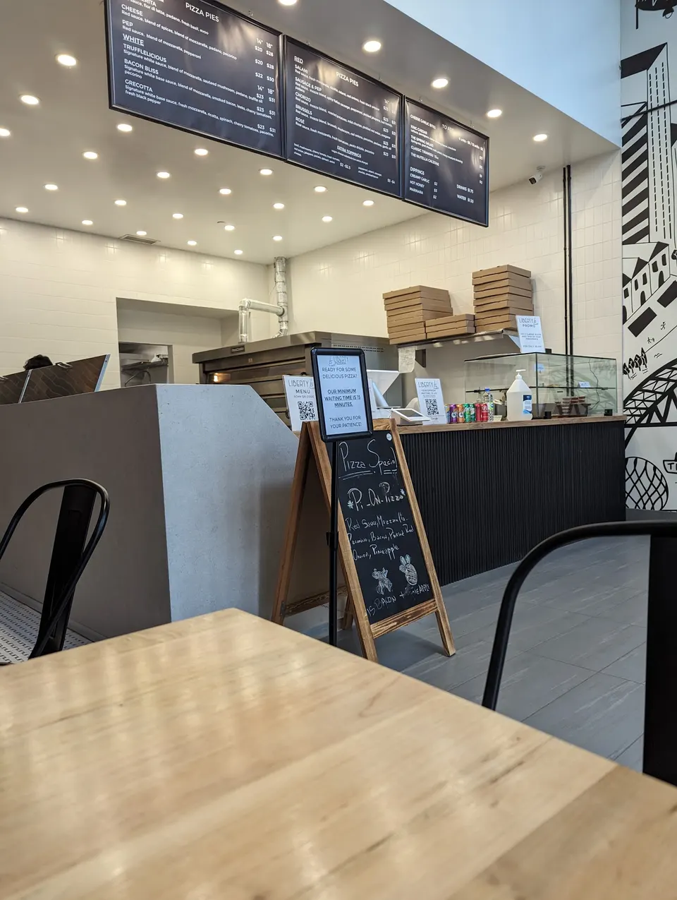 Interior view of Liberty Pizzeria showing a wooden table in the foreground, a black chalkboard menu sign, the order counter, and overhead menu boards.