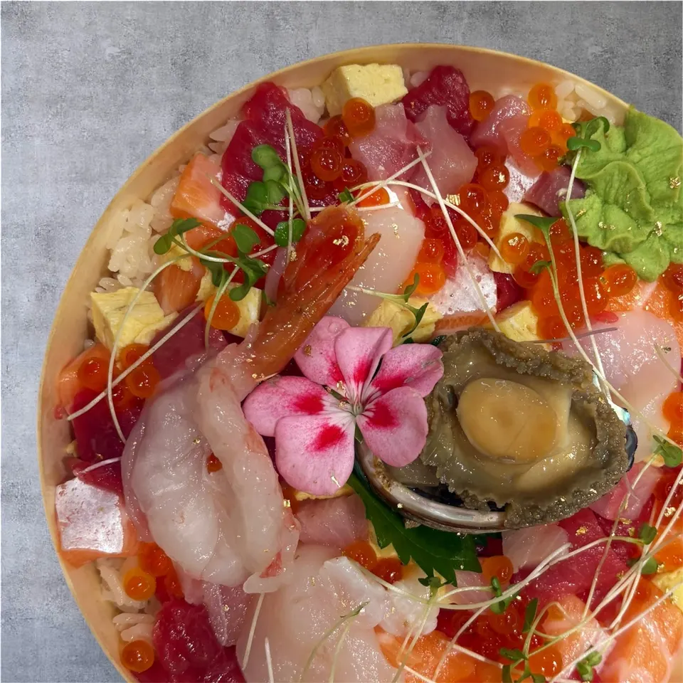 A close-up, top-down view of a colorful chirashi sushi bowl in a round wooden container, featuring raw fish, shrimp, abalone, salmon roe, and a pink flower garnish.
