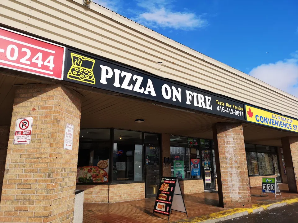 Exterior storefront of Pizza On Fire, featuring a black sign with white lettering, a yellow pizza box logo, and a brick pillar facade under a covered walkway.