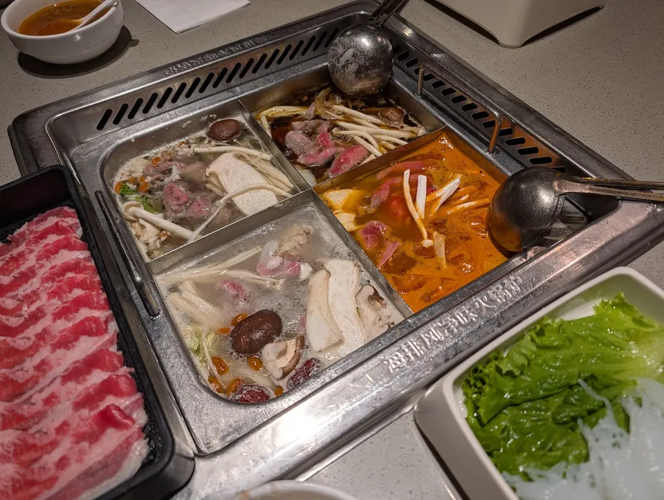 A four-compartment hot pot simmering with different broths, mushrooms, and meats, accompanied by sliced raw beef, fresh lettuce, and glass noodles on a dining table.