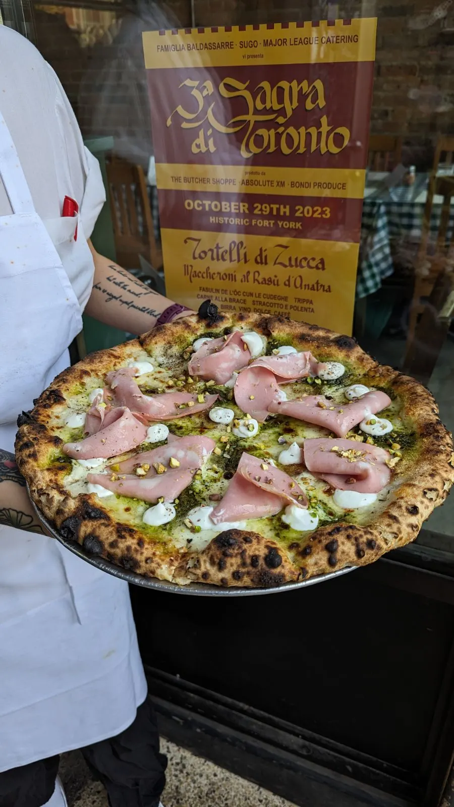 A person in a white apron holding a freshly baked Neapolitan-style pizza topped with mortadella, dollops of white cheese, pistachio pesto, and crushed pistachios, with an event poster in the background.