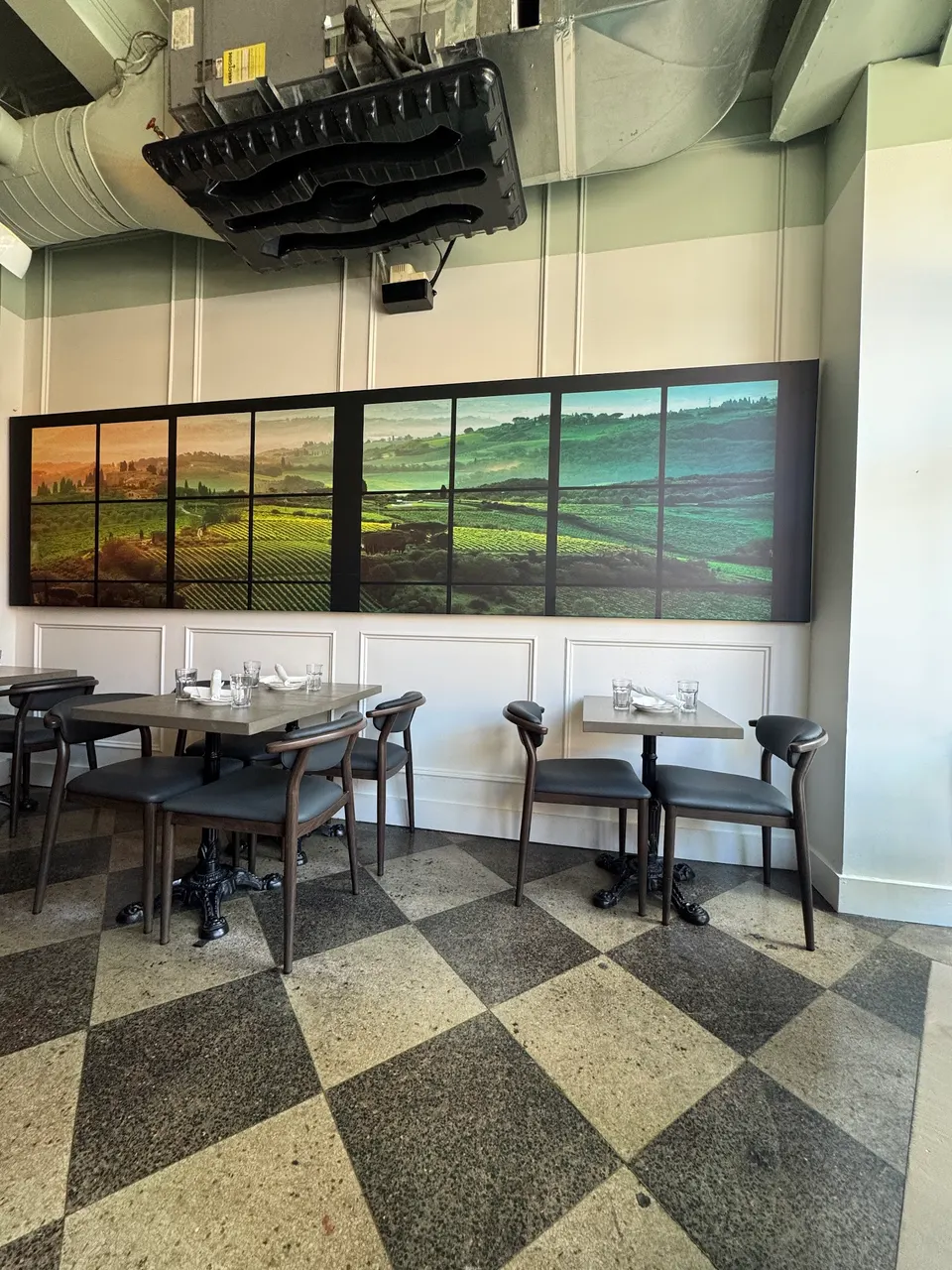 A dining area at Nodo Junction featuring a black and white checkered floor, wooden tables set with glasses, dark grey chairs, and a large panoramic landscape painting of rolling green hills on the wall.