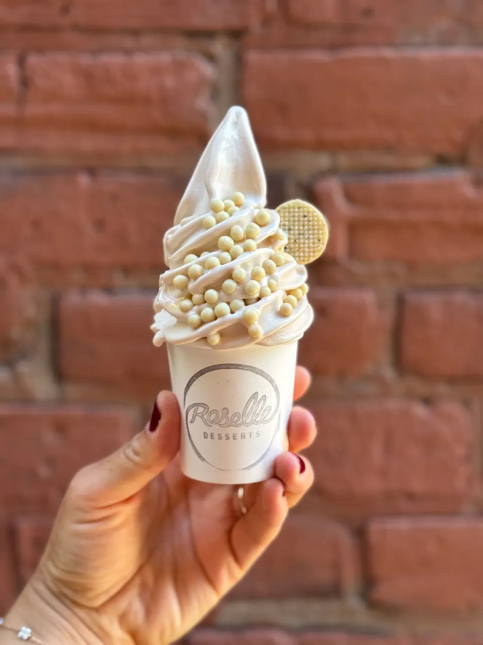 A hand holding a small white cup of soft-serve ice cream topped with white chocolate pearls and a small round wafer, with a brick wall background.