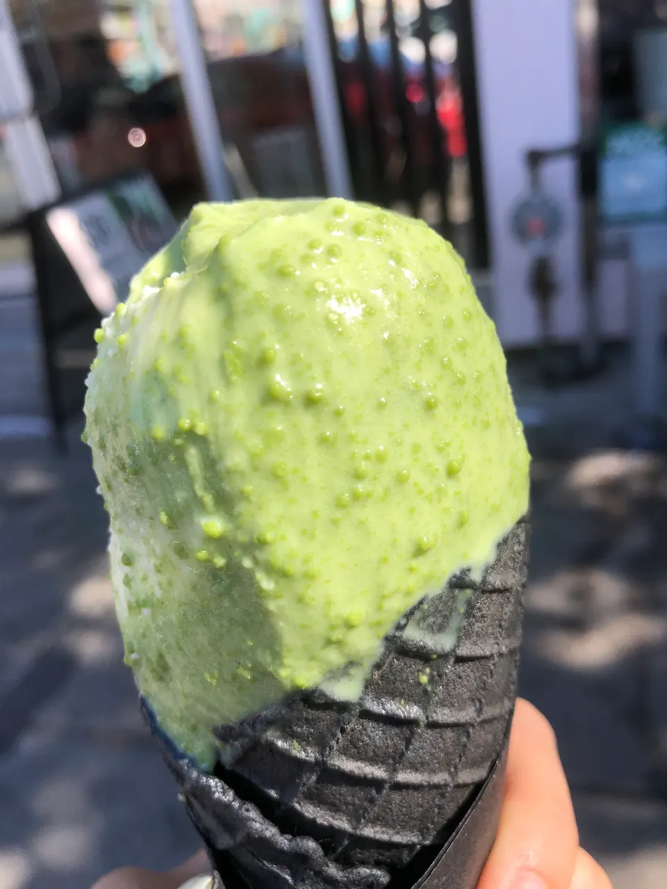 A scoop of bright green matcha ice cream with a textured surface, served in a black charcoal waffle cone held by a hand outdoors.