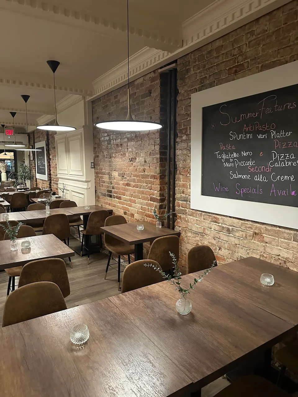 Cozy dining room at Pizza e Pazzi featuring dark wood tables, brown upholstered chairs, exposed brick walls, and a large chalkboard menu displaying summer features.